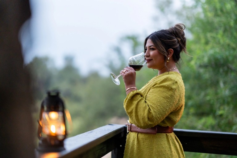 Serene Evening at Silvan Safari A woman in a yellow dress enjoys a glass of wine while standing on a wooden deck, with a lantern lit beside her, surrounded by greenery in the background.