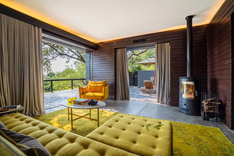 Cassia Suite Lounge Area The image showcases the stylish lounge area of the Cassia Suite, featuring cozy yellow furniture, large windows offering a view of nature, and a fireplace, creating a warm and inviting atmosphere.