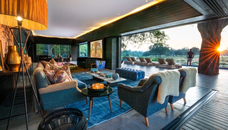 The image showcases a beautifully designed lounge area at Silvan Safari, featuring stylish seating arrangements, a coffee table, and a picturesque view overlooking the sunset and outdoor seating.
