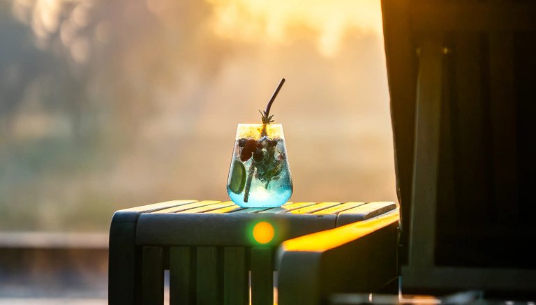 A beautifully styled cocktail glass filled with vibrant blue liquid and garnished with fruits sits on a wooden table, bathed in golden sunset light.