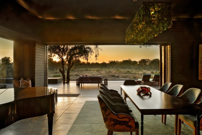 An elegant dining area with a polished black piano, a long wooden table, and chairs featuring leopard patterns, showcasing a beautiful sunset and outdoor scenery through large glass windows.