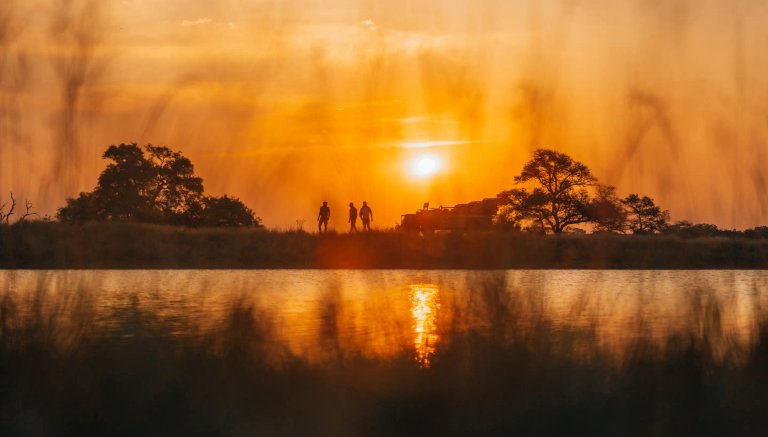 A picturesque sunset scene over a water body with silhouettes of three people and a safari vehicle in the background, framed by tall grass and trees.