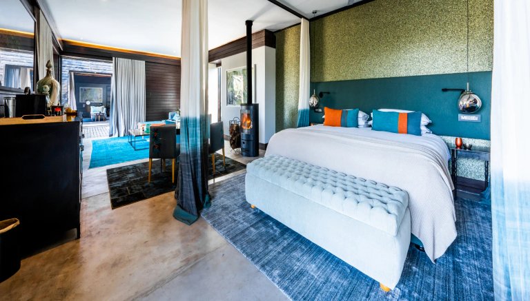 The image showcases a beautifully designed room in Silvan Safari, featuring a large bed with colorful cushions, a cozy seating area, and elegant decor. Natural light fills the space from the open patio area visible through the curtains.