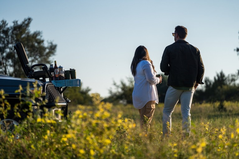 A couple enjoys a beautiful sunset view while standing near a safari vehicle, with drinks in hand, amidst a field of wildflowers.