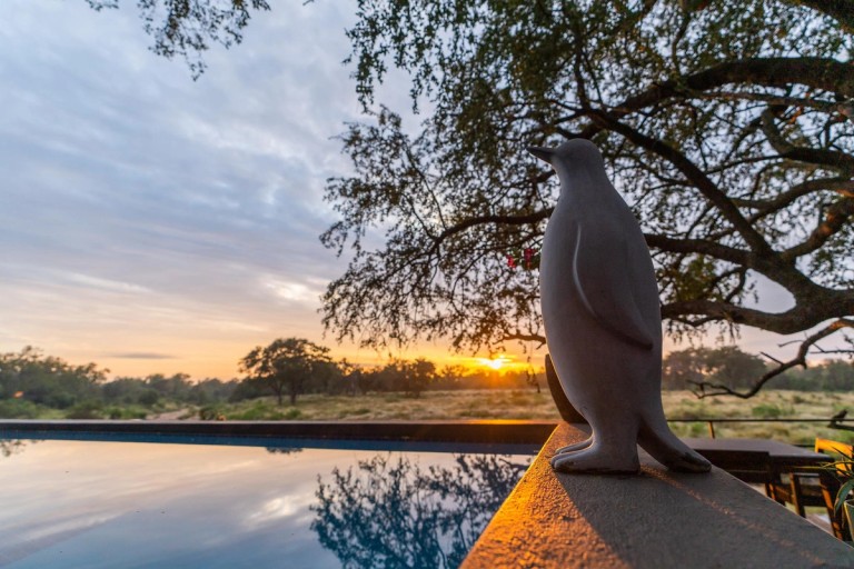 Penguin Sculpture Silhouette at Sunset A decorative penguin sculpture stands on a ledge, silhouetted against a vibrant sunset sky reflecting on the water of a pool. The surrounding landscape is lush with trees.