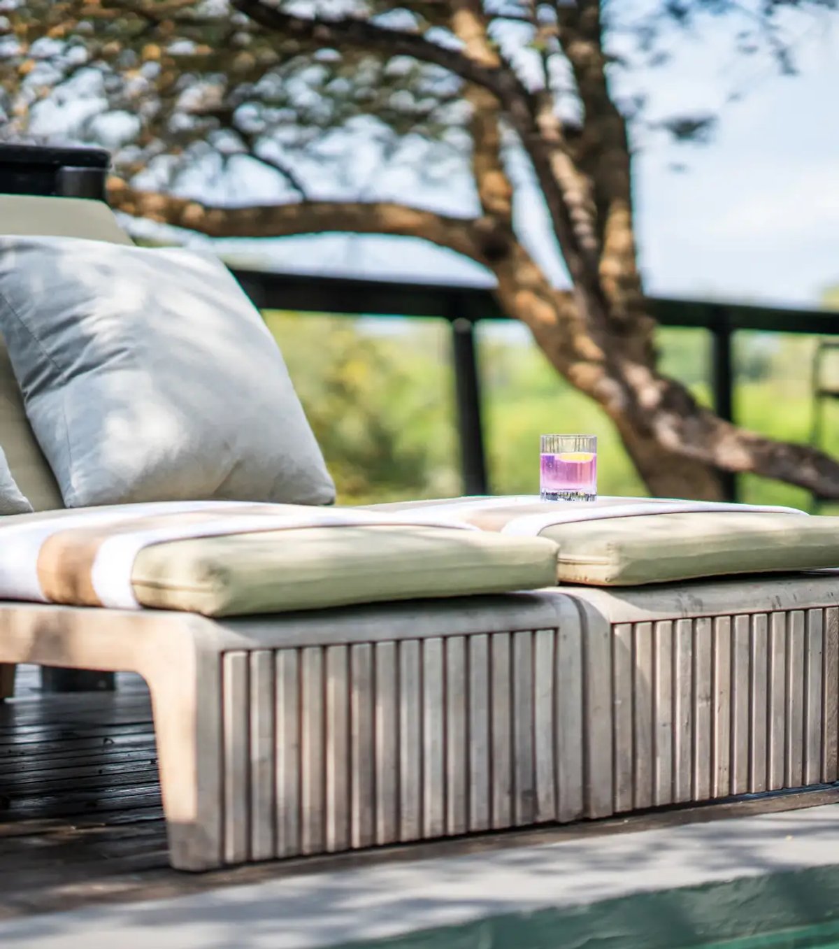 A relaxing lounge area featuring a wooden chaise lounge with soft cushions and a colorful drink placed on it, surrounded by nature.