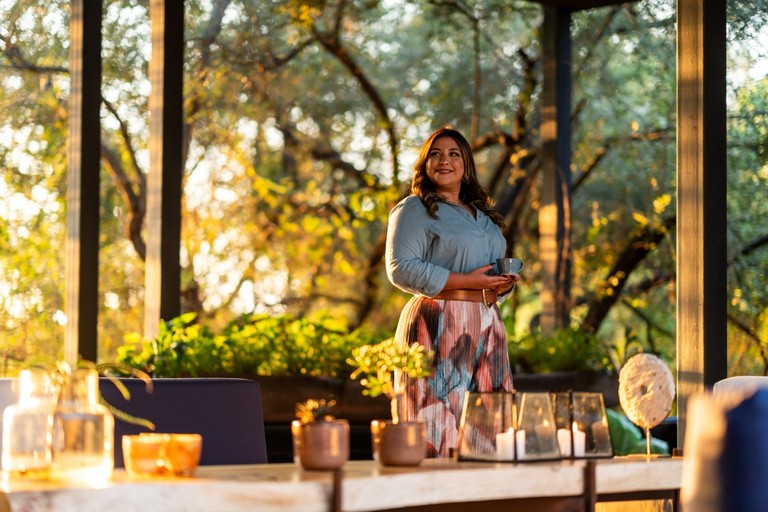 A smiling woman stands in a well-decorated space surrounded by greenery, holding a cup, capturing the inviting atmosphere of Silvan Safari.