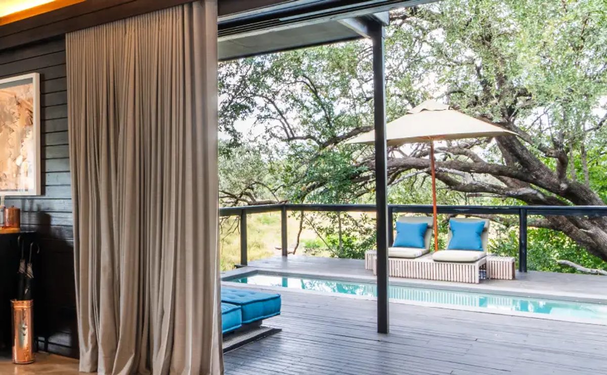 The image showcases a beautifully designed room at Silvan Safari Lodge, featuring a modern aesthetic with large curtains and a view of an outdoor pool area surrounded by trees.