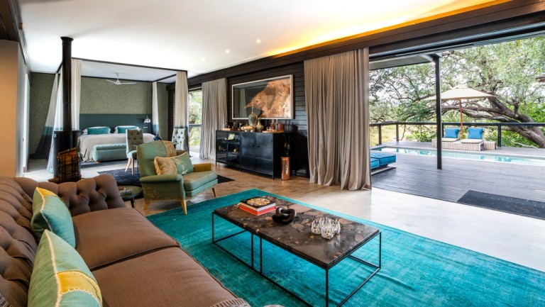 Silvan Safari Lounge Room The image showcases a beautifully designed lounge area in Silvan Safari, featuring comfortable furniture, a cozy fireplace, and large windows that open to a serene outdoor space with a swimming pool.