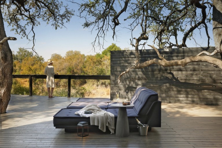 A tranquil relaxation area at Silvan Safari, featuring a lounging sofa and scenic views of the surrounding wilderness. A woman stands at the edge, enjoying a peaceful moment in nature.