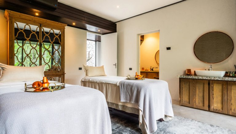 The image shows a beautifully designed spa room with two massage beds, adorned with plush blankets and decorative items. Natural light filters in through the large windows, creating a serene ambiance.
