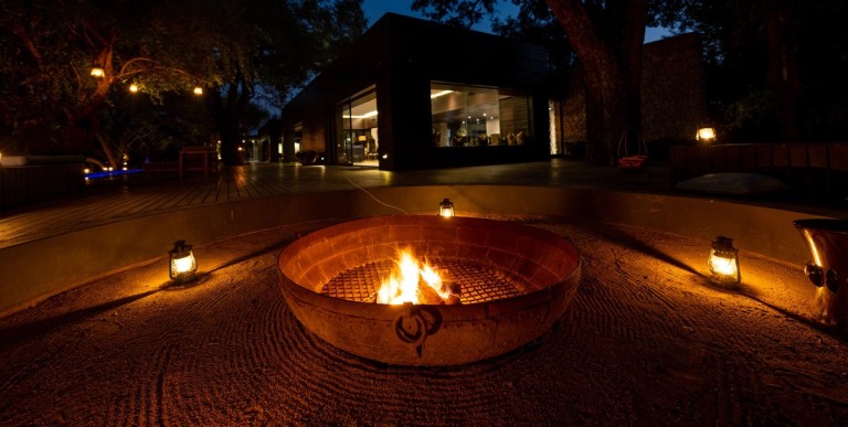 A cozy evening scene featuring a fire pit surrounded by glowing lanterns at Silvan Safari. The ambiance is warm, with a modern lodge in the background.