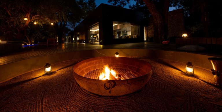 Silvan Safari Evening Fire Pit A cozy evening scene featuring a fire pit surrounded by glowing lanterns at Silvan Safari. The ambiance is warm, with a modern lodge in the background.