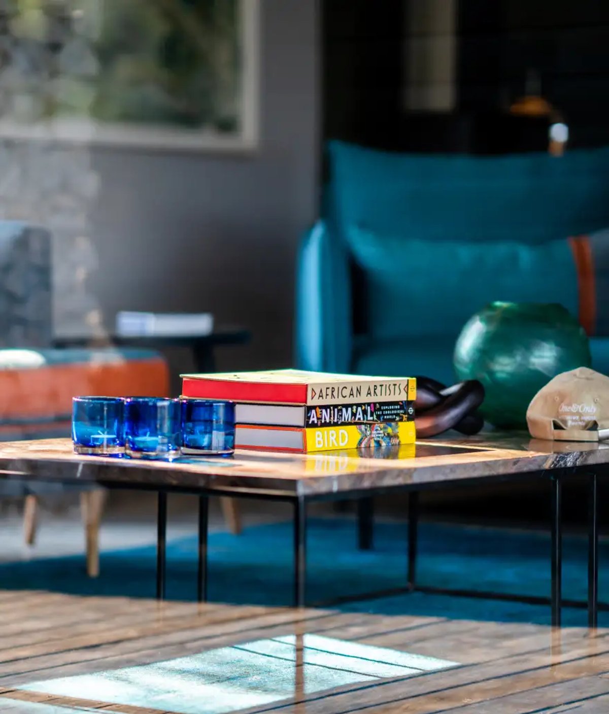 The image showcases a comfortable interior space featuring a table with three blue glass cups, several stacked books including titles related to African artists and animals, and decorative items like a green vase and a hat.