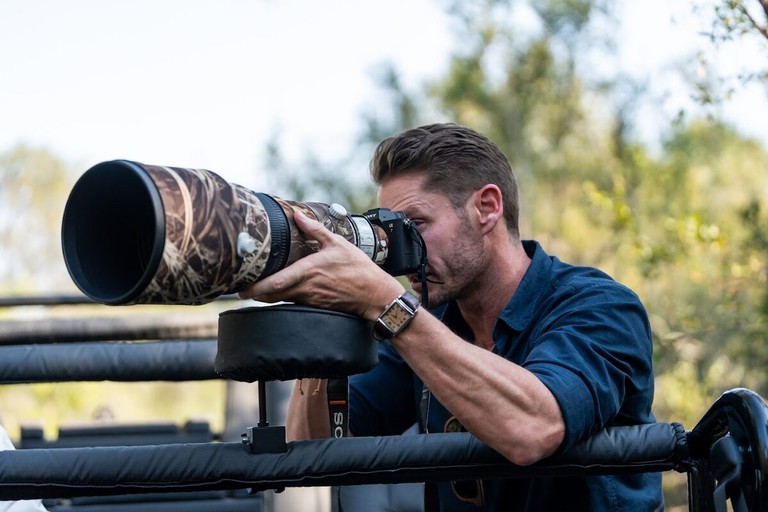 Silvan Safari Wildlife Photographer A wildlife photographer is focusing intently on his camera equipped with a large telephoto lens, ready to capture the beauty of nature during a safari at Silvan Safari.