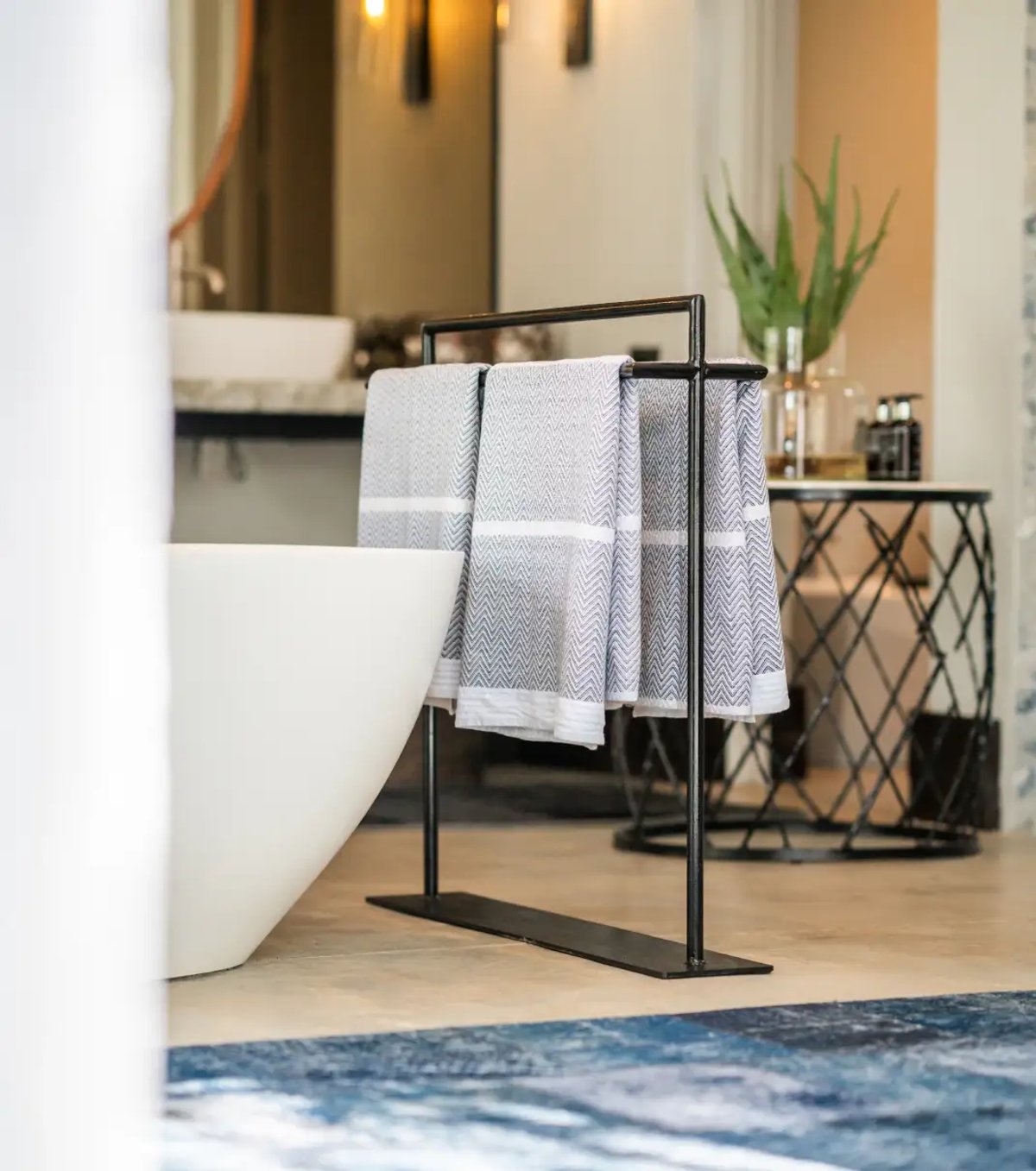 The image showcases a modern bathroom area with a freestanding white bathtub, neatly arranged towels on a towel rack, and a stylish decor including a plant and decorative items on a side table.