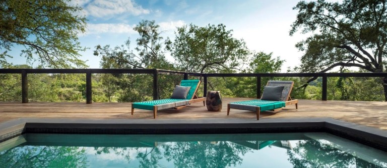 A serene outdoor lounge area at Silvan Safari, featuring two teal loungers beside a pool with a scenic view of lush trees and a clear sky.