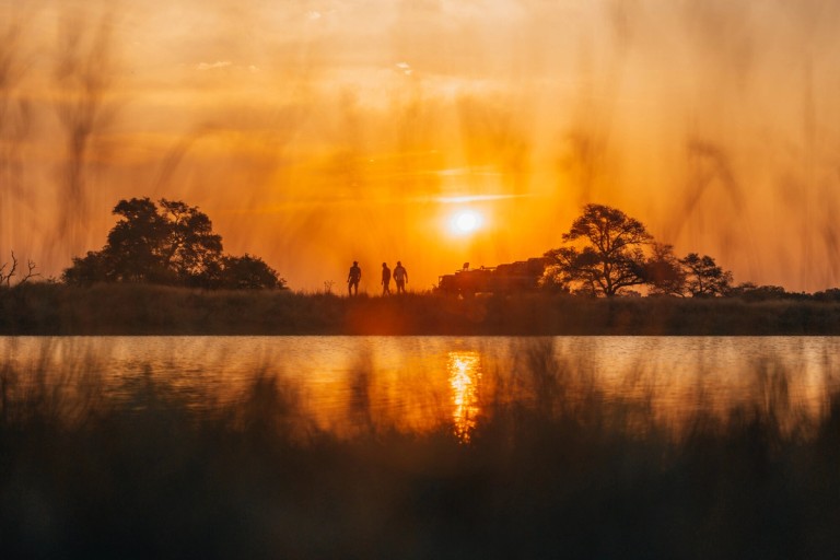 A stunning sunset landscape at Silvan Safari, showcasing silhouettes of people against a vibrant orange sky, reflecting on the water.