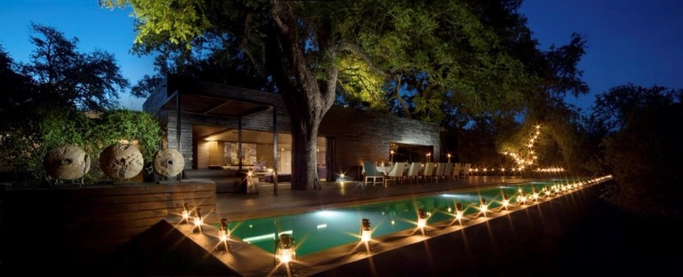 A serene evening view of the Silvan Safari Lodge, featuring an illuminated pool, dining area, and ambient lighting among lush surroundings.