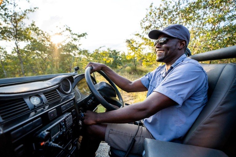 A safari guide is smiling while driving an open-top safari vehicle in a lush, green environment, suggesting a joyful experience in the wilderness.