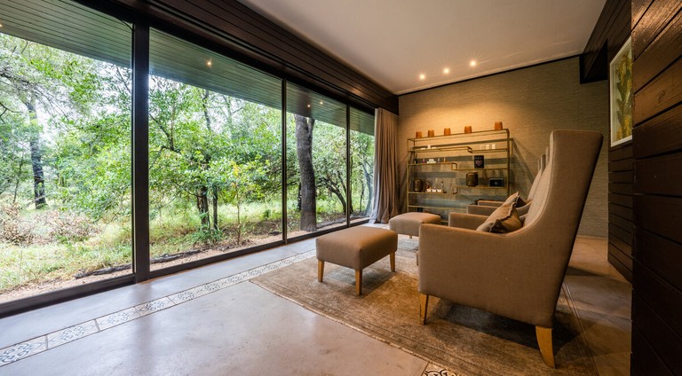 The image shows a cozy lounge area at Silvan Safari Lodge featuring comfortable seating and a view of the lush surroundings through large windows.