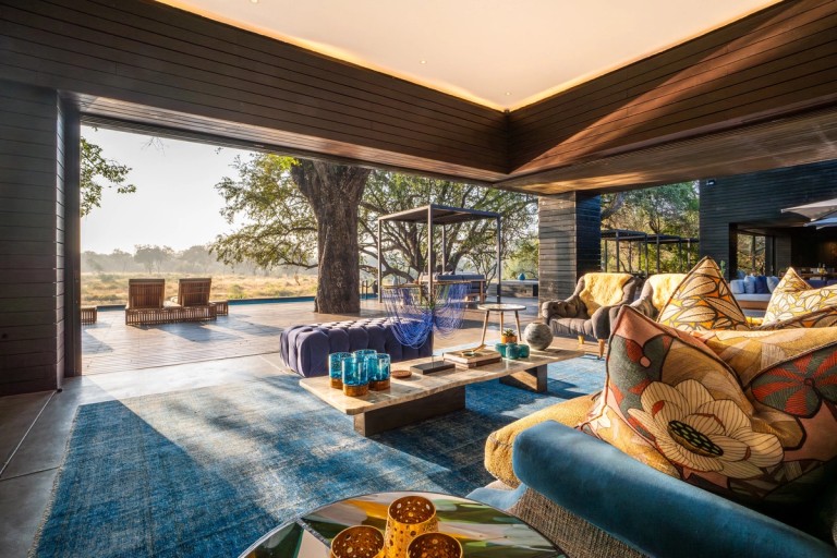 A beautifully designed lounge area at Silvan Safari Lodge featuring elegant furniture and decor. The scene opens to a stunning view of the outdoor landscape, including a large tree and seating on the deck.